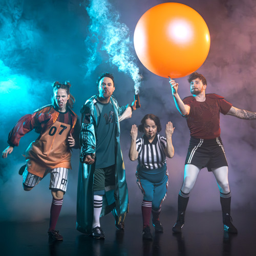 Skådespelarna står med fotbollskläder. Killen till höger dribblar med en stor orange boll.