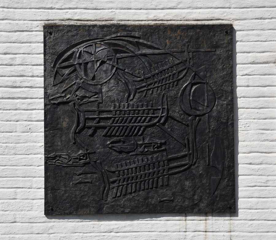 Relief i järn med olika figurer och symboler. Är monterad mot en vit tegelvägg. 